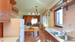 A kitchen with wooden cabinets, a white refrigerator, a dining table with chairs, a chandelier, and a window with curtains.