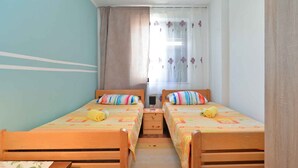 Una habitació amb dos llits de fusta, cadascun amb un llençol i almoades de colors. Hi ha una finestra amb cortines transparents i un dibuix floral, i una taula petita entre els llits.