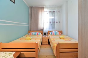 Ein Zimmer mit zwei Holzbetten, jedes mit farbenfroher Bettdecke und Kissen. Ein Fenster mit transparenten Vorhängen und floralem Muster, und ein kleiner Tisch zwischen den Betten.