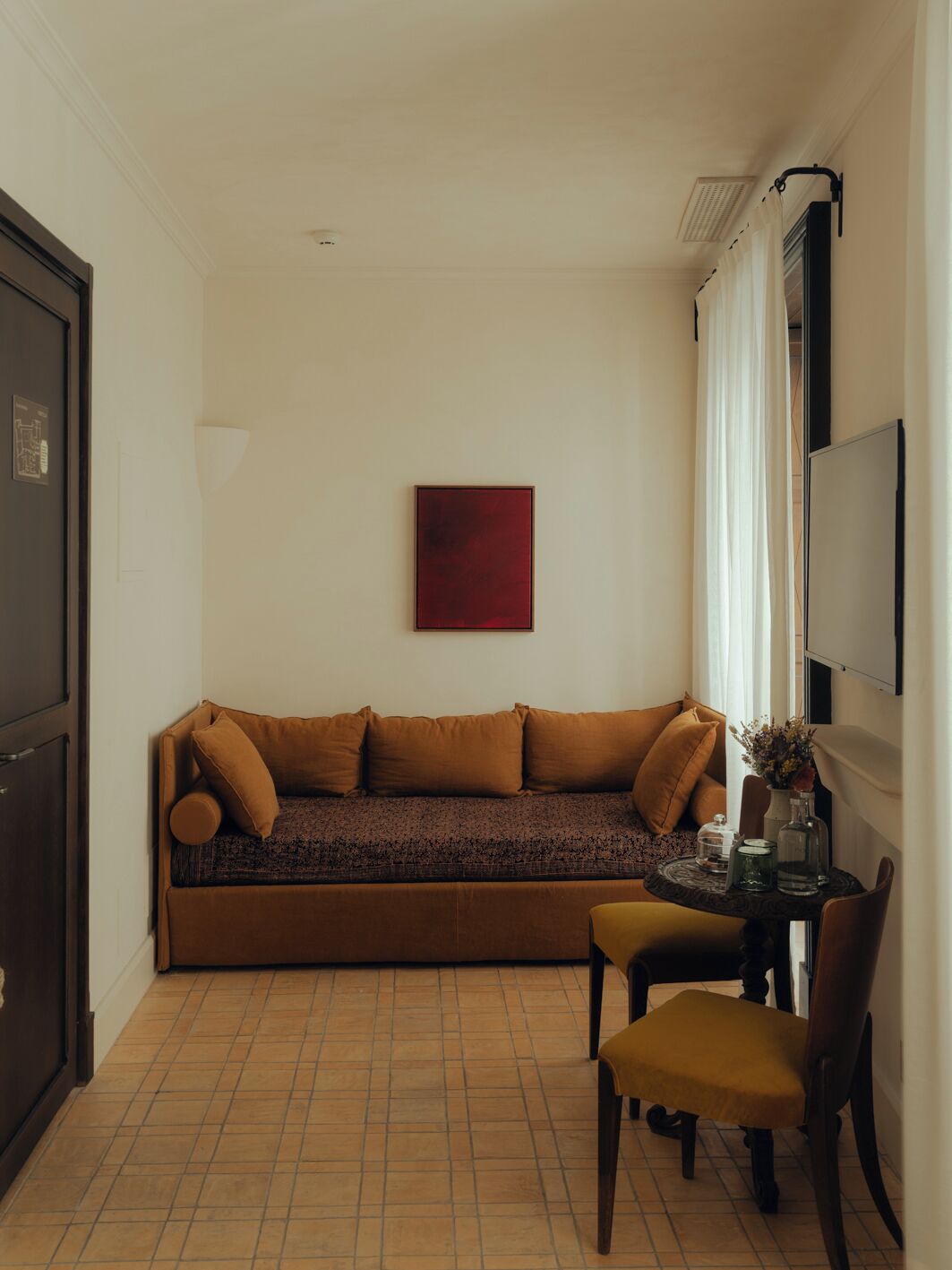 Una habitación pequeña y acogedora con un sofá marrón, una mesa redonda con una silla, una obra de arte roja en la pared y una ventana con cortinas blancas.