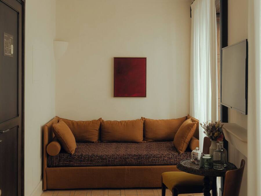 Una habitación pequeña y acogedora con un sofá marrón, una mesa redonda con una silla, una obra de arte roja en la pared y una ventana con cortinas blancas.