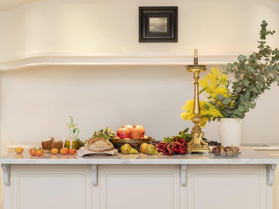 A kitchen counter with various fruits, vegetables, a loaf of bread, a bottle, and a decorative candle holder.