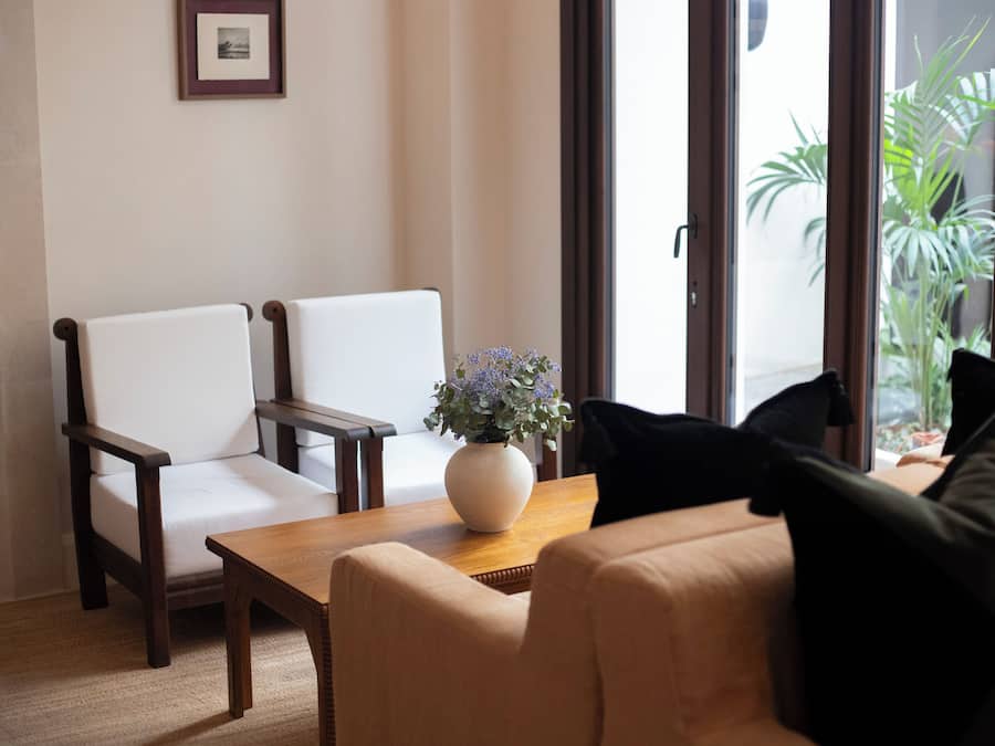 Una habitación con dos sillas blancas, una mesa de madera con un florero y un sofá color crema.