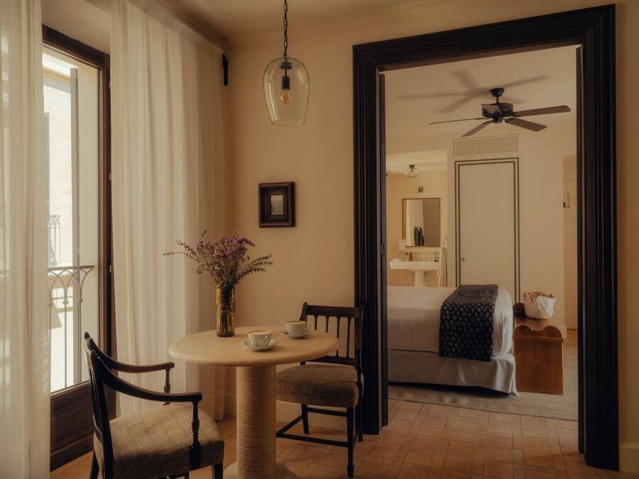 Una habitación con una mesa redonda, dos sillas, un florero con flores, un ventilador de techo y vistas a un baño a través de una puerta abierta.