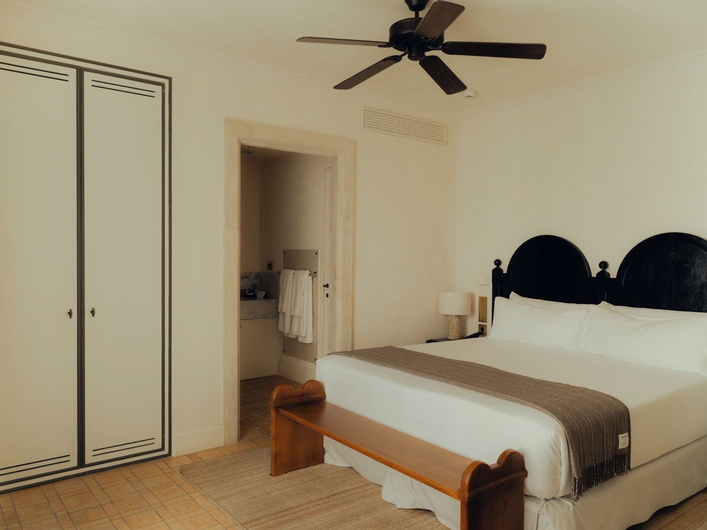 Un dormitorio con cama, mesita de noche, ventilador de techo, armario y un baño visible a través de una puerta abierta.