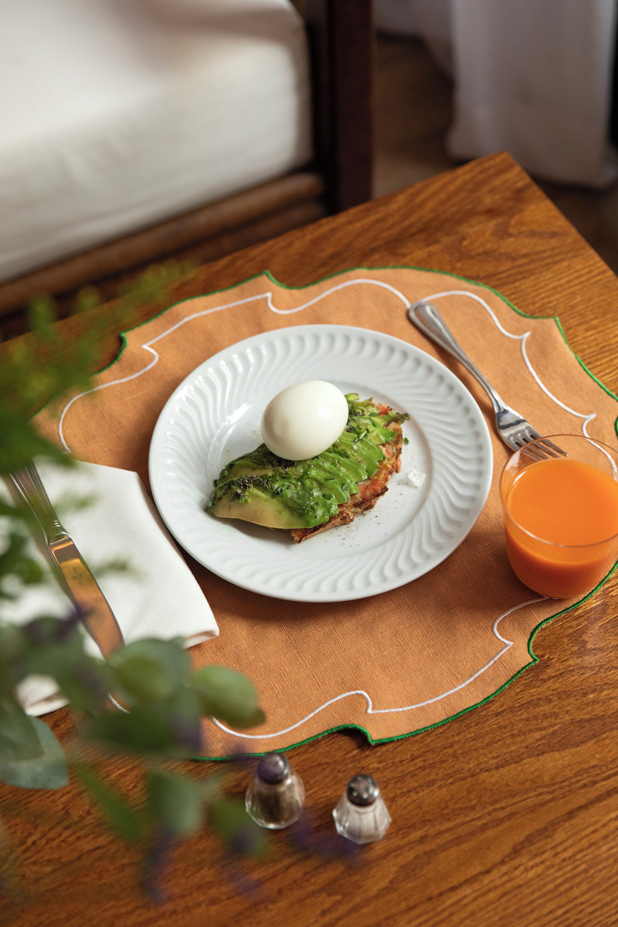 Un plato con un sándwich y un huevo escalfado sobre un plato blanco, un vaso de jugo de naranja y una servilleta sobre una mesa de madera.