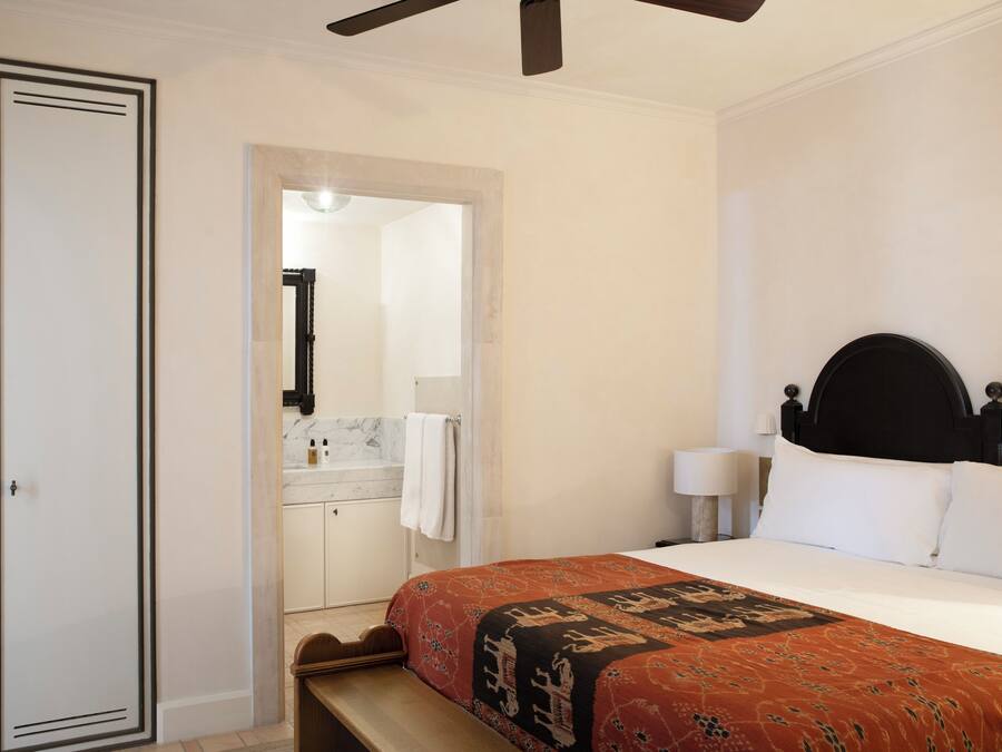 Un dormitorio con cama, mesita de noche, escritorio, silla, armario y baño visible a través de una puerta abierta.
