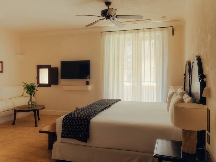 Habitación de hotel con cama, ventilador de techo, televisor, ventana con cortinas translúcidas, mesita de noche y lámpara.
