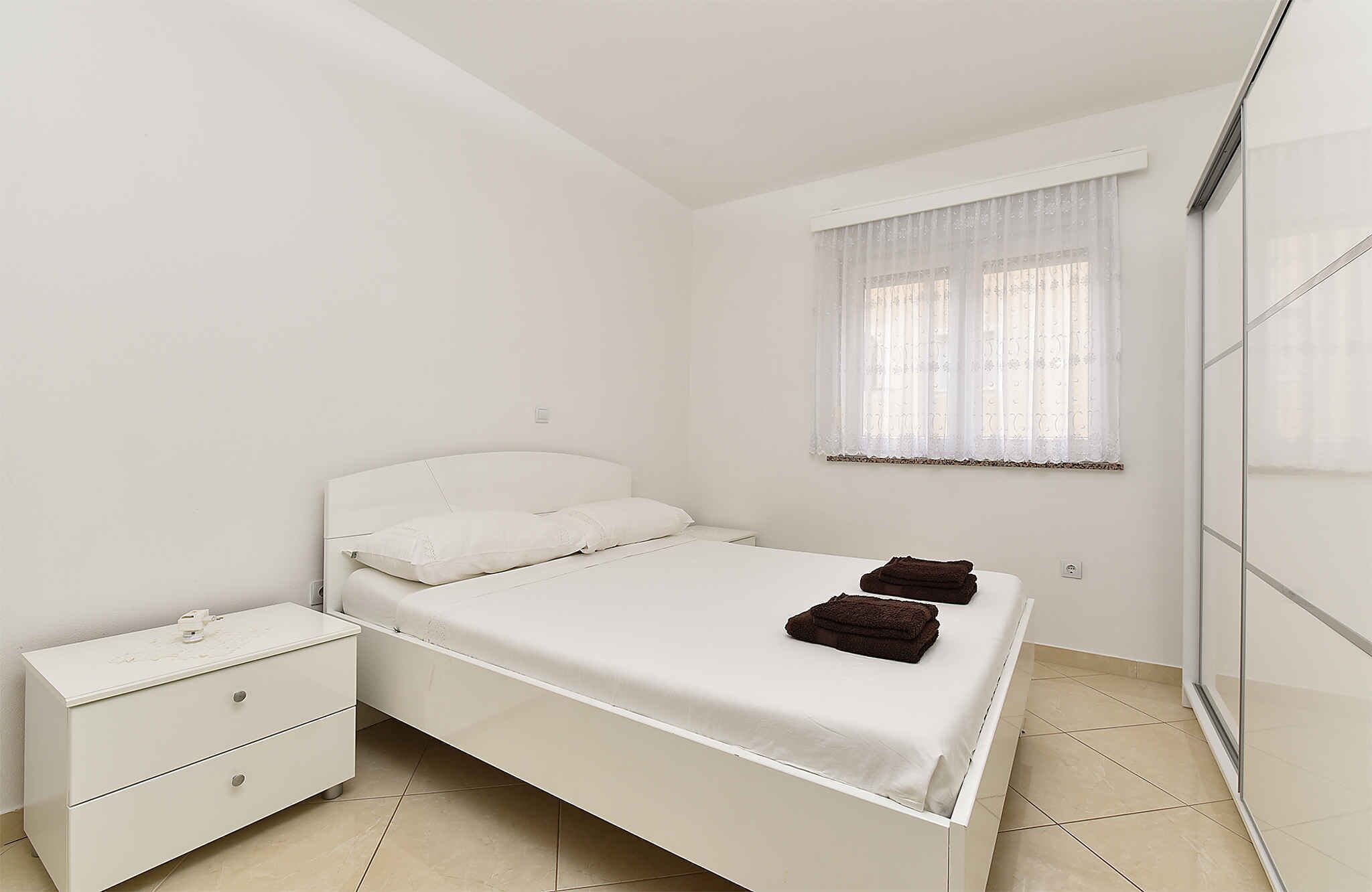 Un dormitorio blanco con una cama, una mesita de noche, una ventana con cortinas translúcidas y un separador de vidrio.