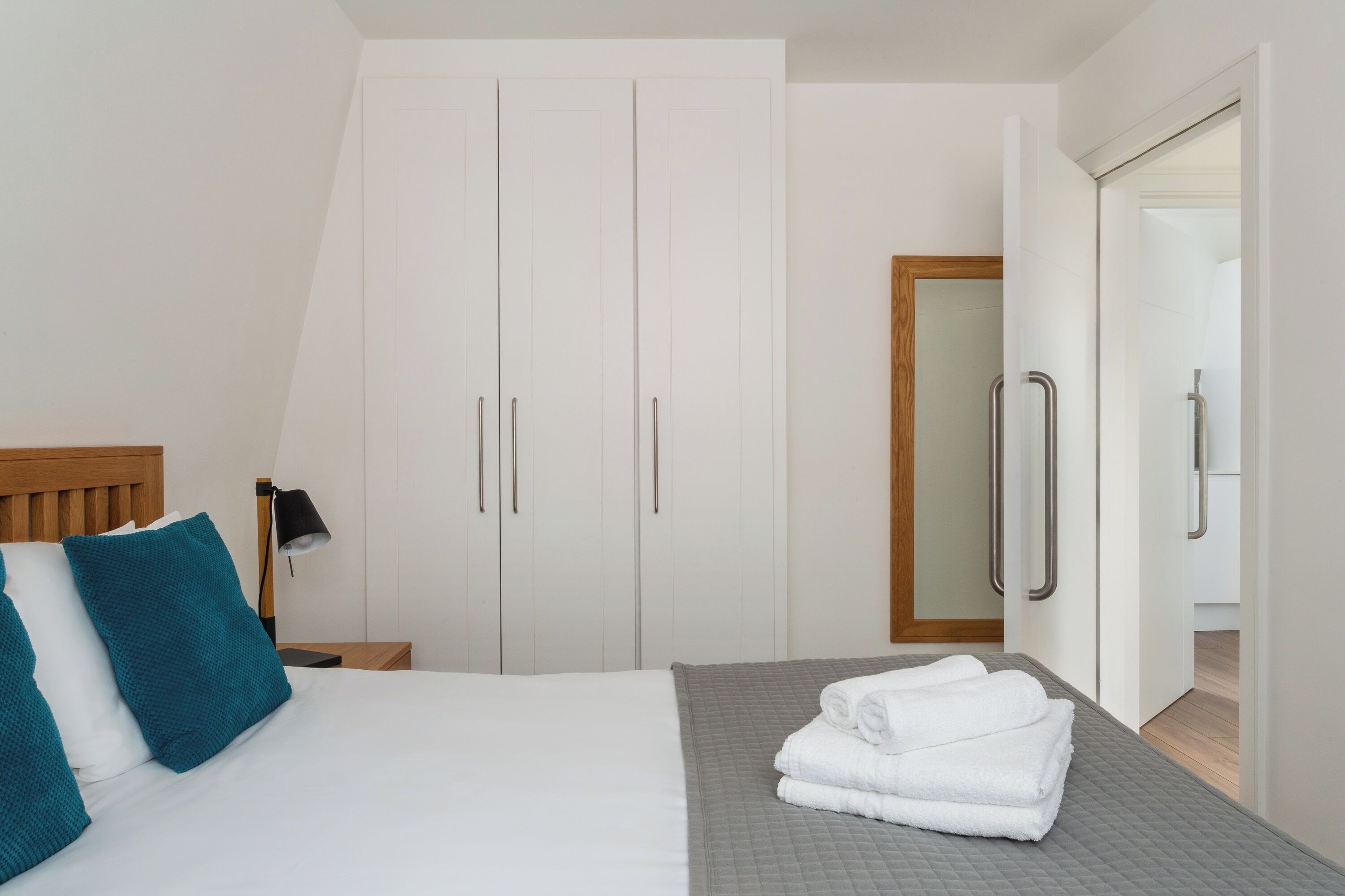 A bedroom with a bed, white curtains, a wooden headboard, a nightstand, a lamp, and a wardrobe.