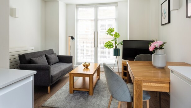 Ein modernes Wohnzimmer mit grauem Sofa, Holztisch und einem Esstisch mit Stühlen. Es gibt ein großes Fenster mit Jalousien, eine Stehlampe und einen Blumentopf mit Blumen.