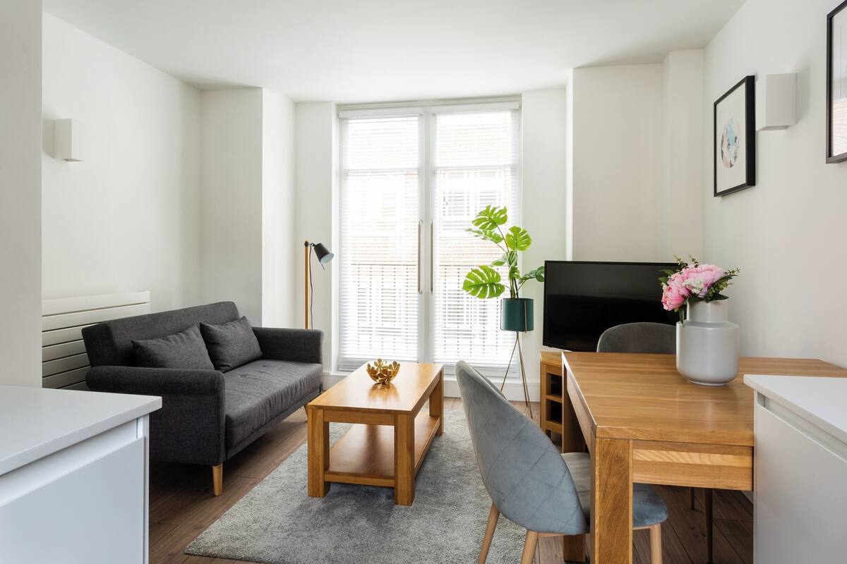 A modern living room with a grey sofa, wooden coffee table, and a dining table with chairs. There is a large window with blinds, a floor lamp, and a potted plant with flowers.
