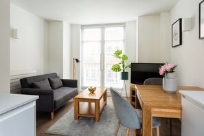 En moderne stue med en grå sofa, et træ sofabord og et spisebord med stole. Der er et stort vindue med persienner, en gulvlampe og en potteplante med blomster.