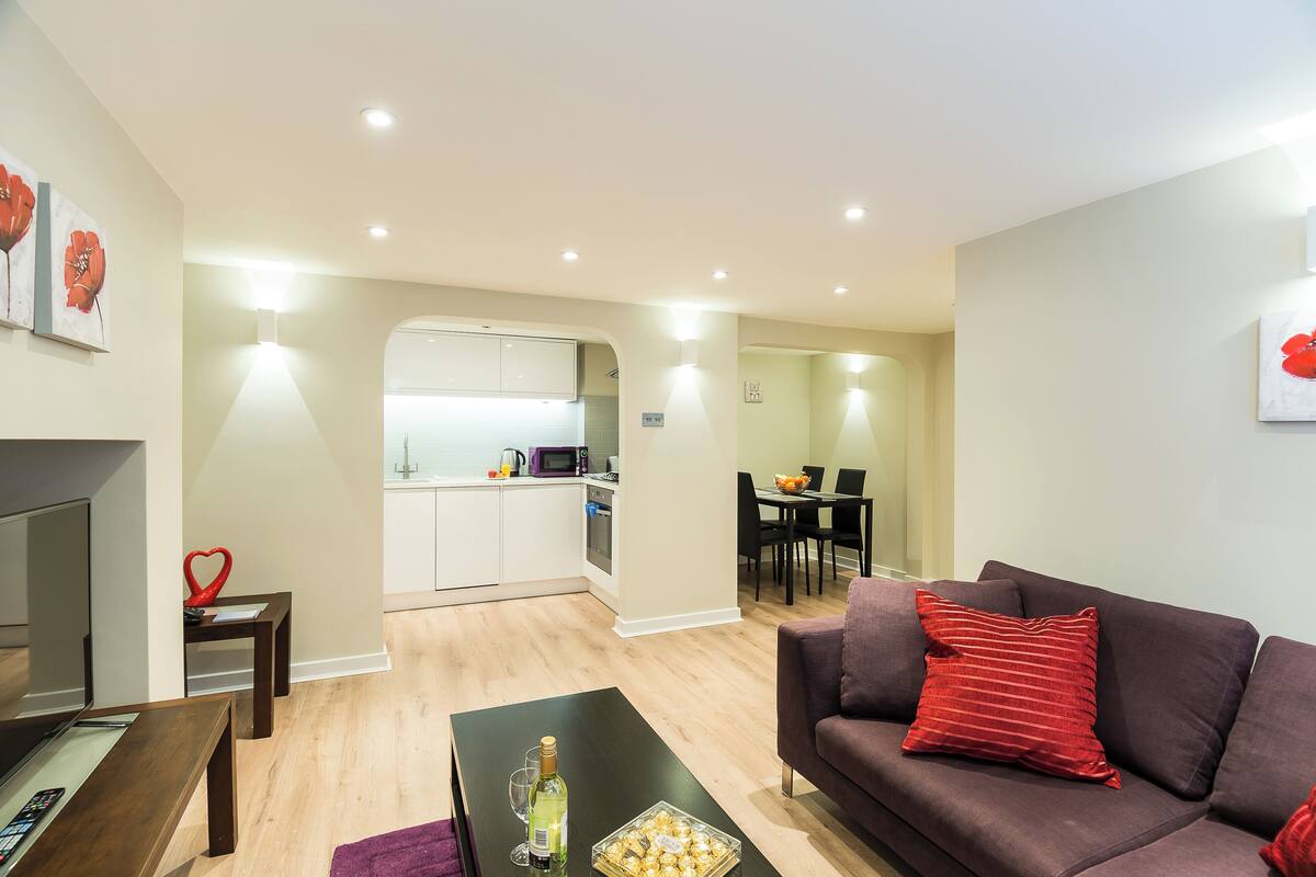A modern living room with a dark brown sofa, a small coffee table, and a TV. A kitchen with white cabinets and a dining area with a table and chairs is visible in the background.
