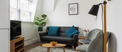 A modern living room with a grey sofa, blue cushions, a round coffee table, a TV on a wooden stand, a floor lamp, and a potted plant.