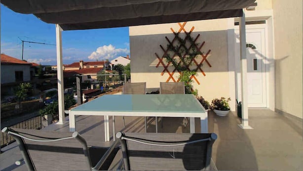 An outdoor patio with a glass-top table and chairs, a decorative wall, and a view of the neighborhood.