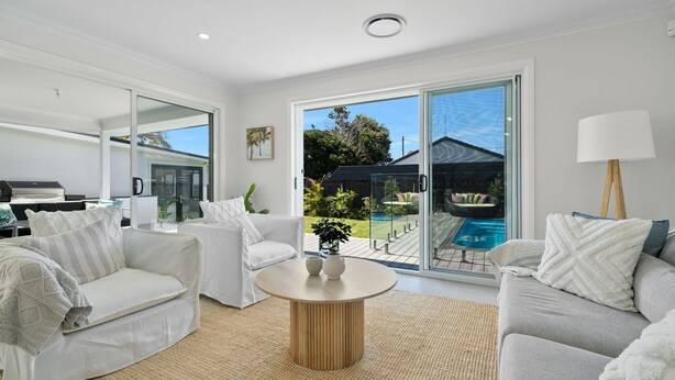 A modern living room with a large sliding glass door leading to a pool area. There are two white sofas, a round wooden coffee table, and a floor lamp.