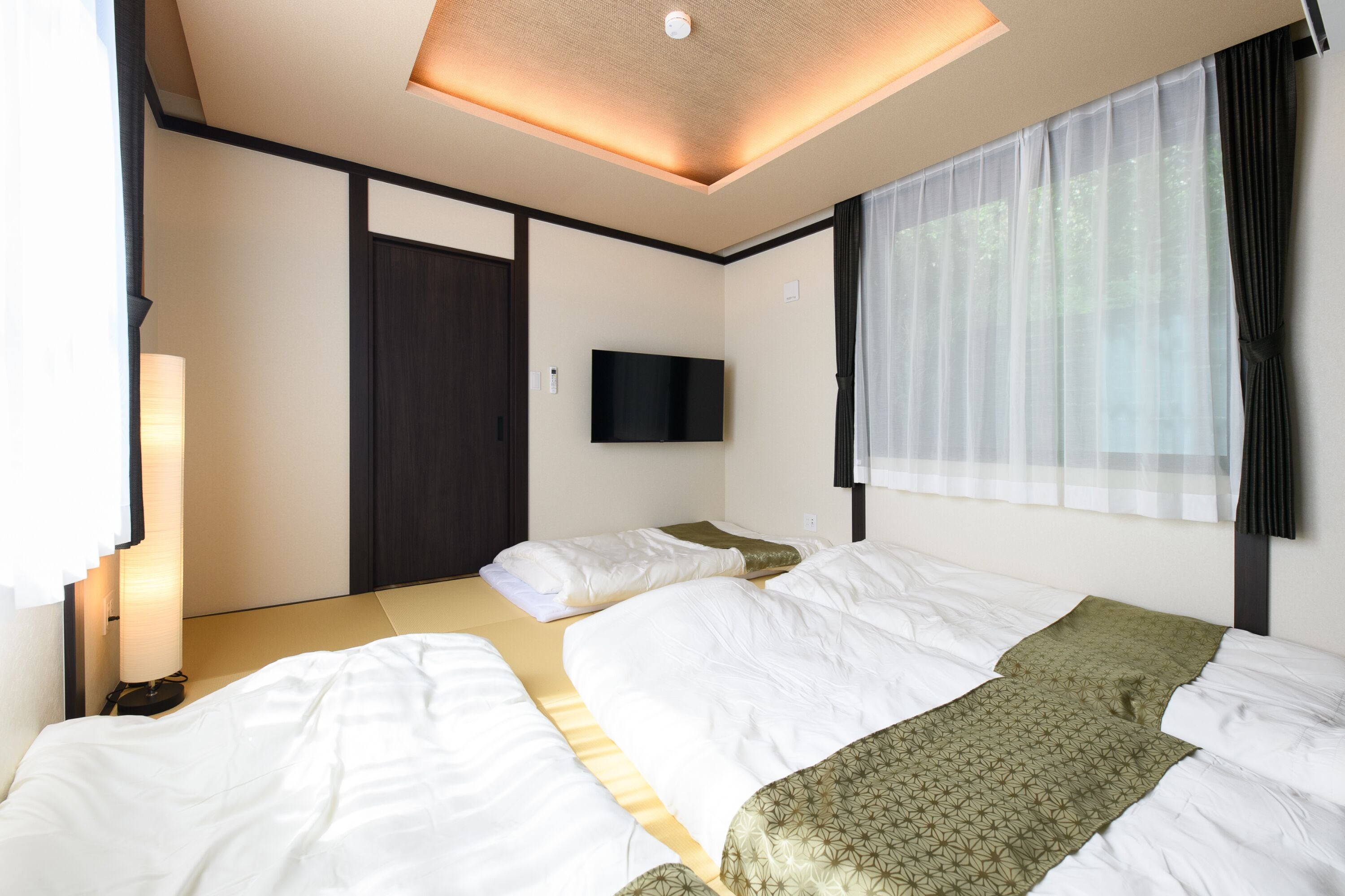 A hotel room with two beds, a flat-screen TV, a window with sheer curtains, and a ceiling light fixture.