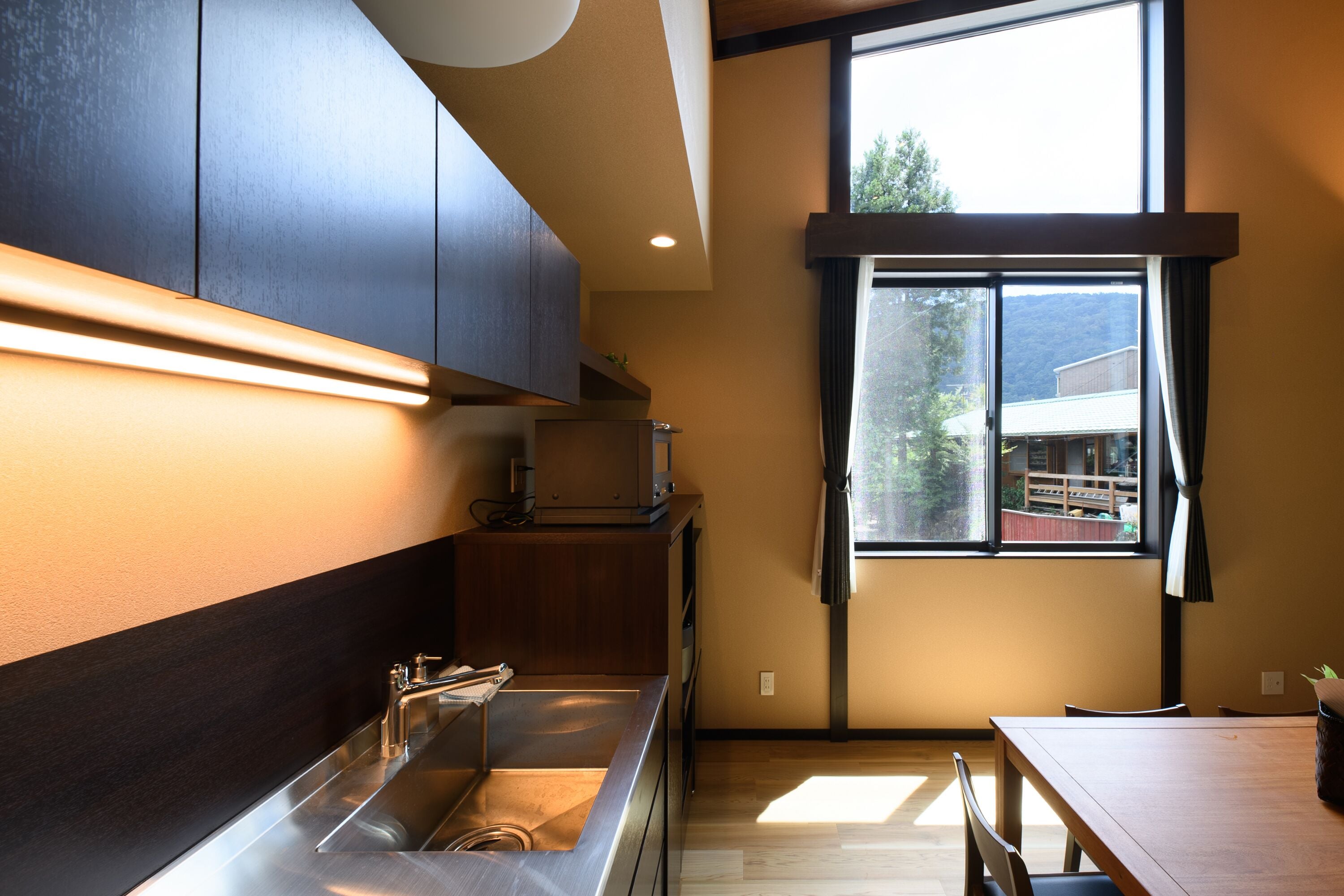 A modern kitchen with a stainless steel sink, wooden cabinets, and a large window offering a view of a building and trees.