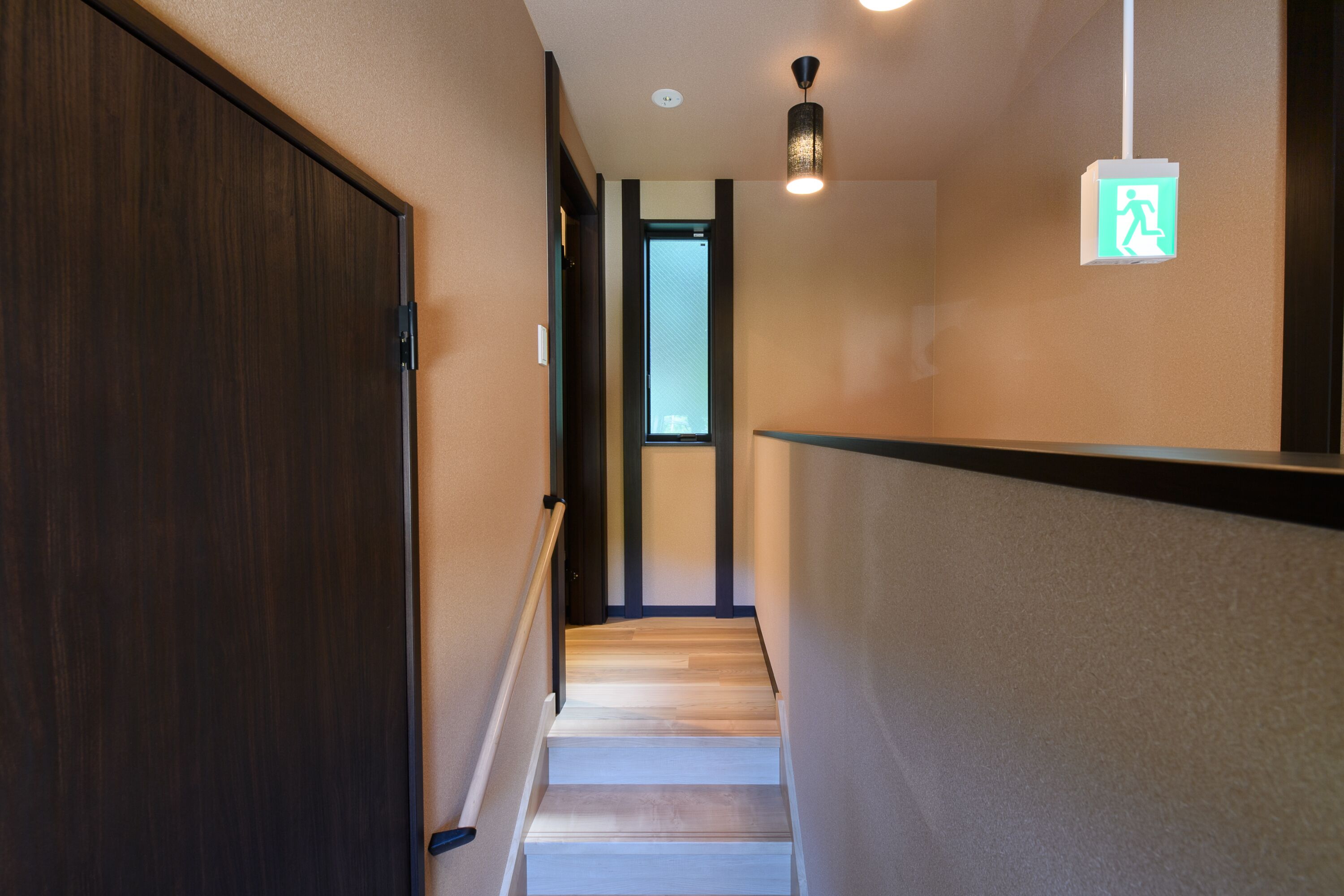 Un couloir étroit, au sol en bois, avec une porte sombre sur la gauche et un mur de couleur claire sur la droite. Une petite fenêtre est visible au fond.