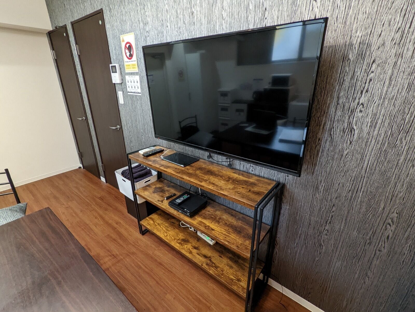 A modern room with a large flat-screen TV mounted on a textured wall, a wooden console table with electronic devices, and a door with a no-smoking sign.