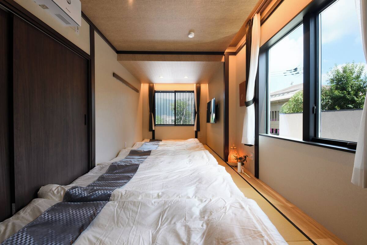 A narrow hotel room with a bed, a window with blinds, a TV mounted on the wall, and a ceiling fan.