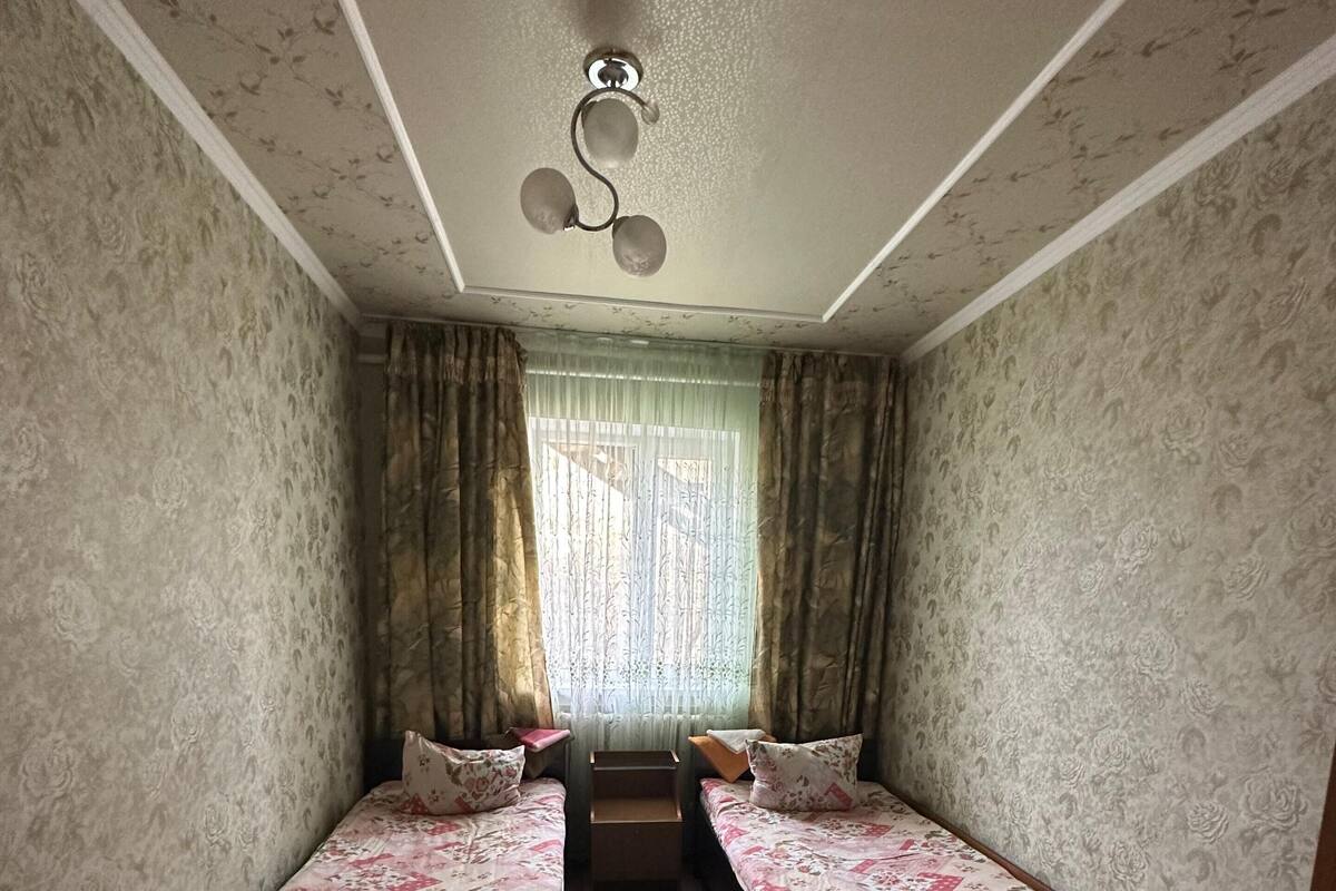 Een kamer met twee eenpersoonsbedden, een klein raam met gordijnen, een plafondlamp en behang met een patroon.