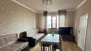 A living room with a sofa, a dining table with chairs, a television, and a window with curtains. - Zhenja and Lyuda's Boarding House (Zhabagly)
