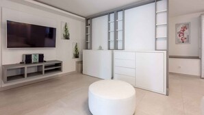 Een moderne woonkamer met een flatscreen televisie, een witte poef en inbouwplanken.