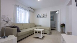 Un salon moderne avec un canapé d’angle beige, une petite table blanche et un ventilateur sur pied.