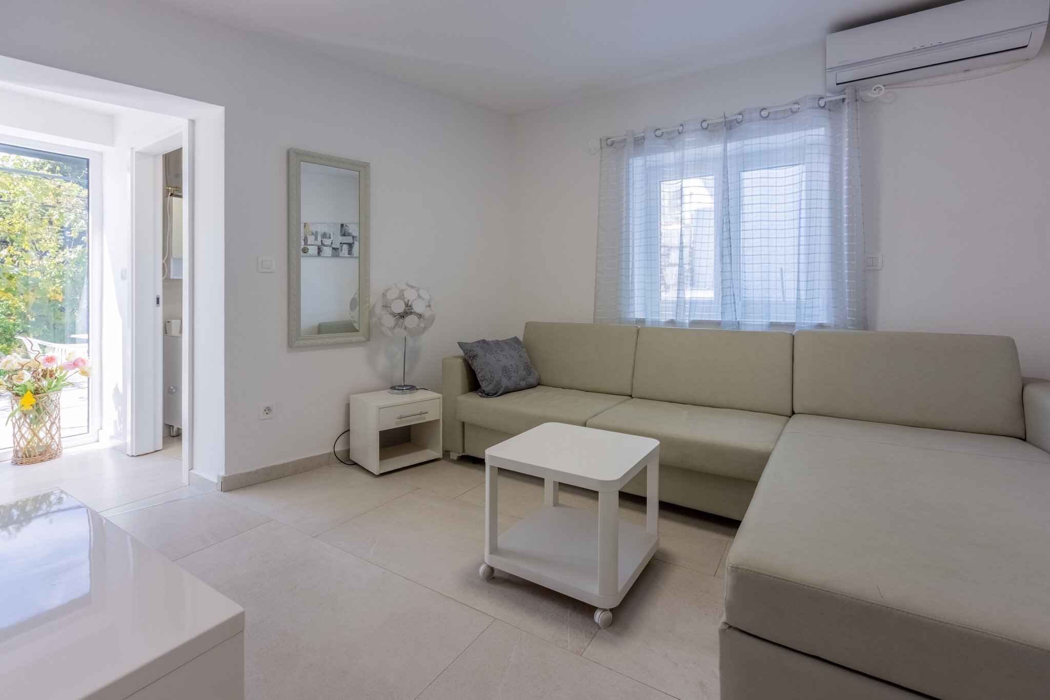 Uma sala de estar moderna com um sofá, uma mesinha branca e uma mesa de cabeceira. Há uma janela com cortinas e um ventilador na mesa de cabeceira.