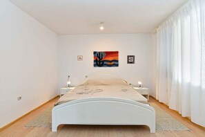 Ein Schlafzimmer mit einem Bett, zwei Nachttischen, einem Schreibtisch und einem Wandbild, sowie einem Fenster mit Vorhängen.