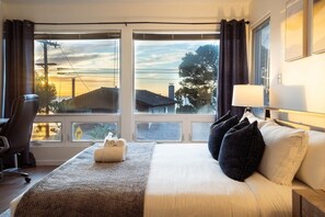 Une chambre d’hôtel avec un grand lit, un bureau et une chaise. Il y a des rideaux aux fenêtres et une lampe sur la table de chevet.