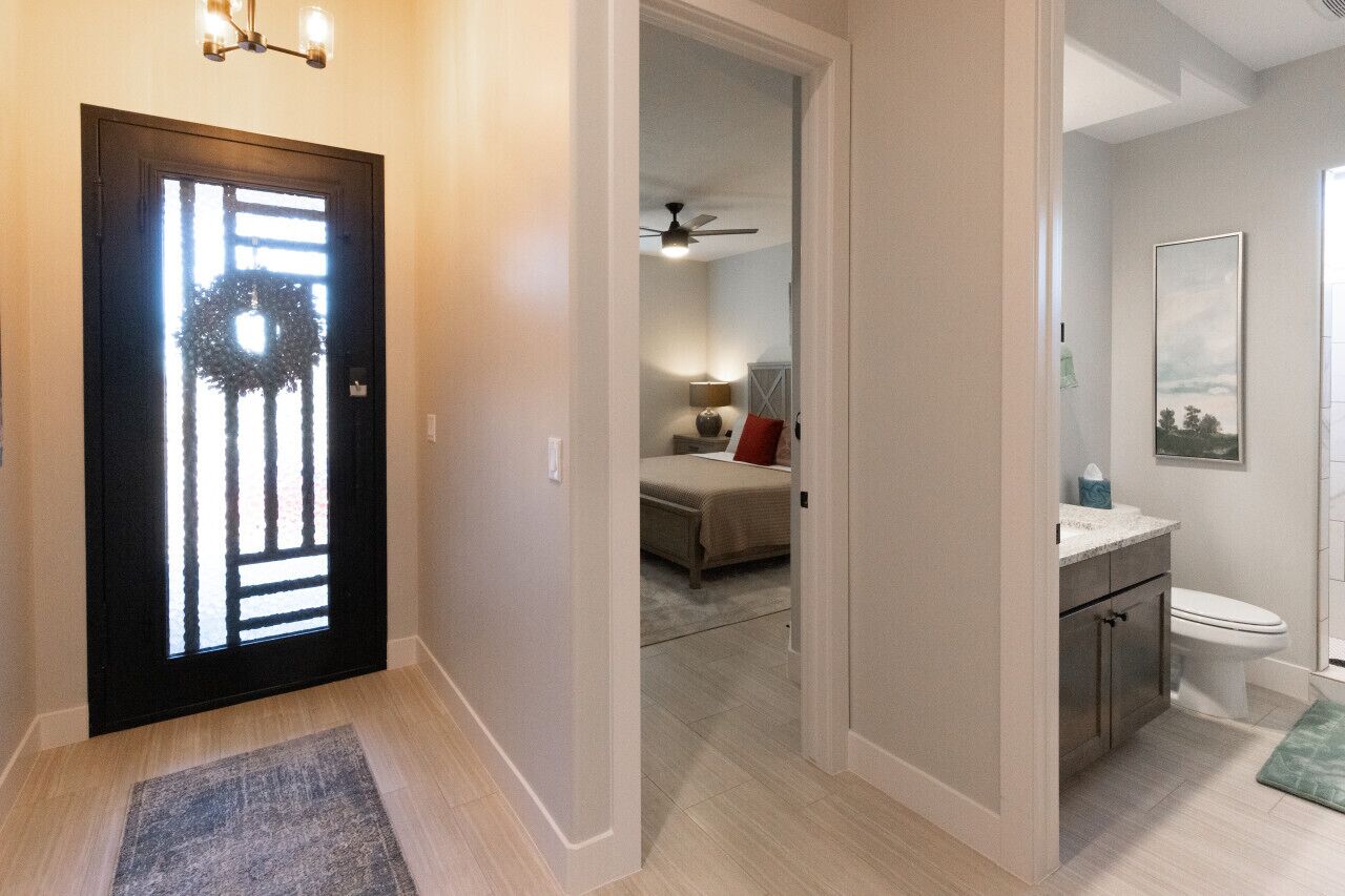 A hallway with a black door featuring a decorative glass panel, a ceiling fan, a bed with a red pillow, and a bathroom with a toilet and a framed picture on the wall.