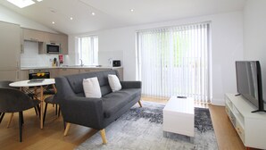 A modern living room with a grey sofa, white coffee table, and a flat-screen TV. There is a dining area with a white table and chairs, and a kitchen with a microwave and oven. - West Finchley by Viridian Apartments (London)