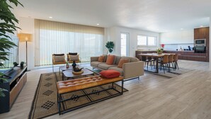 A modern living room with a sofa, ottoman, and coffee table, a dining area with chairs and a table, and a kitchen with appliances.