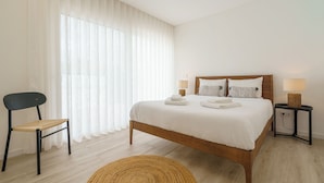 A bedroom with a wooden bed, white bedding, a chair, a bedside table with a lamp, and sheer curtains.