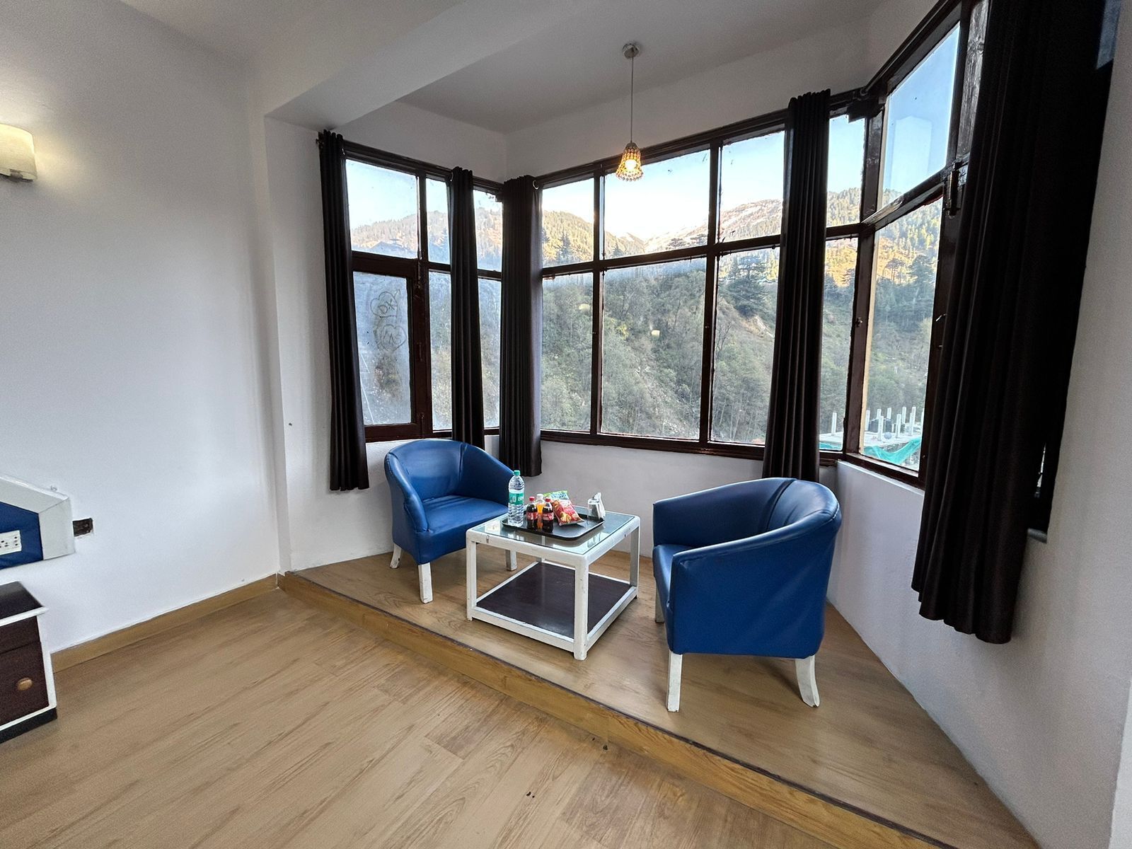 Ein Zimmer mit zwei blauen Stühlen, einem kleinen weißen Tisch und Blick auf eine gebirgige Landschaft durch große Fenster.