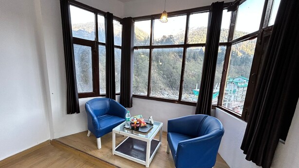 A room with two blue chairs, a small white coffee table, and a view of mountains through large windows. - Hotel Manali Palace (Manali)