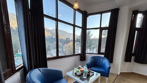 A room with two blue chairs, a glass coffee table with snacks, and a view of a mountainous landscape through large windows. - Hotel Manali Palace (Manali)