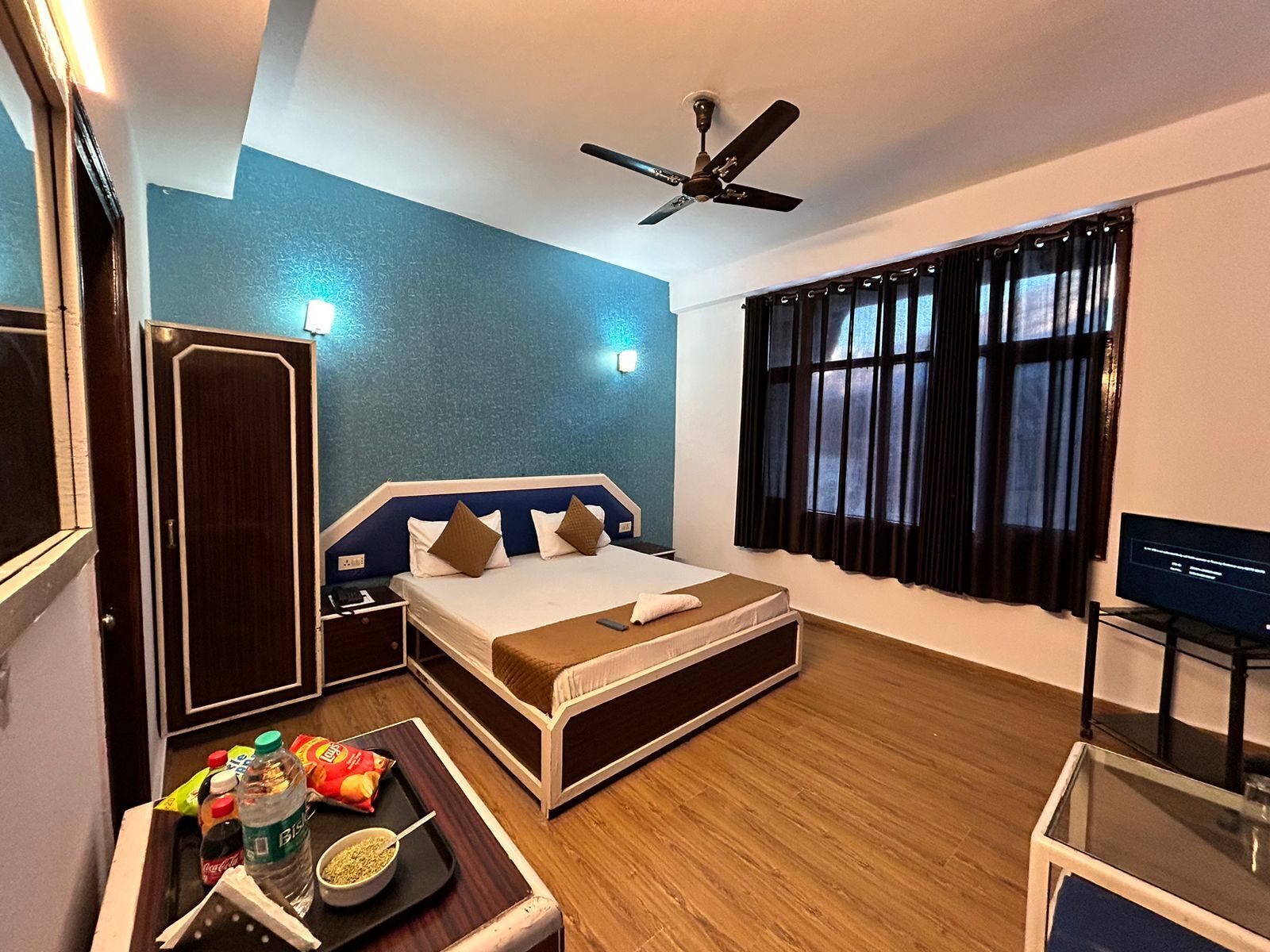 A hotel room with a bed, a desk with a computer, a ceiling fan, a window with curtains, and a door.
