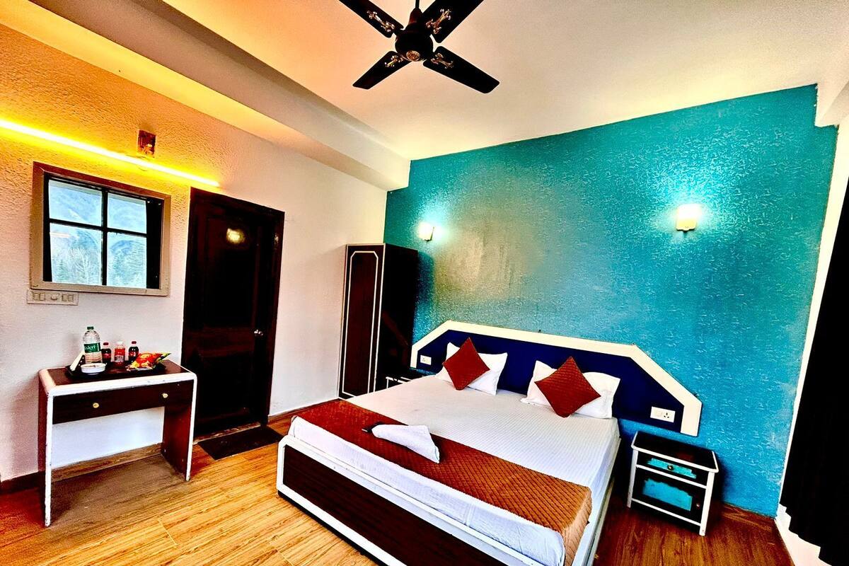 A hotel room with a bed, bedside table, and a desk with a chair. There is a window, a door, and a ceiling fan.