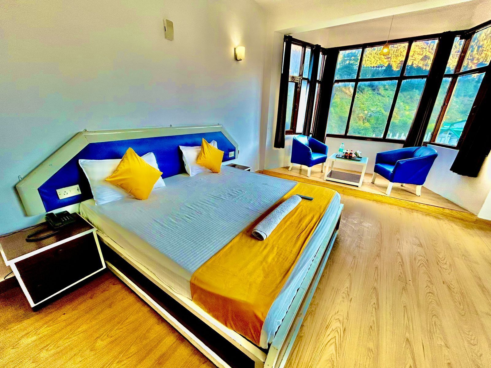 A modern bedroom with a large bed, two blue armchairs, a small table, and a view of greenery through large windows.