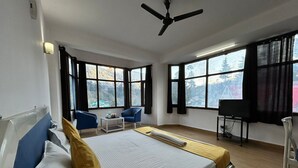 A hotel room with a bed, a desk, two chairs, and large windows offering a view of the outdoors. - Hotel Manali Palace (Manali)