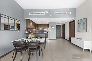 A modern dining area with a long table, four chairs, and a vase of flowers. A kitchen is visible in the background with wooden cabinets and a tiled backsplash. - WelHome - Greenery Beautiful Apartment Onyx Tower 2 (Dubai)