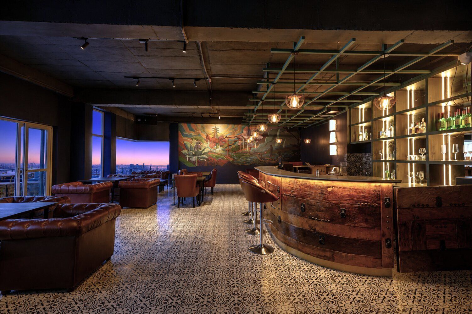 Eine Bar mit einer gebogenen Holztheke, Ledersitzgelegenheiten und Blick auf die Stadt durch große Fenster.