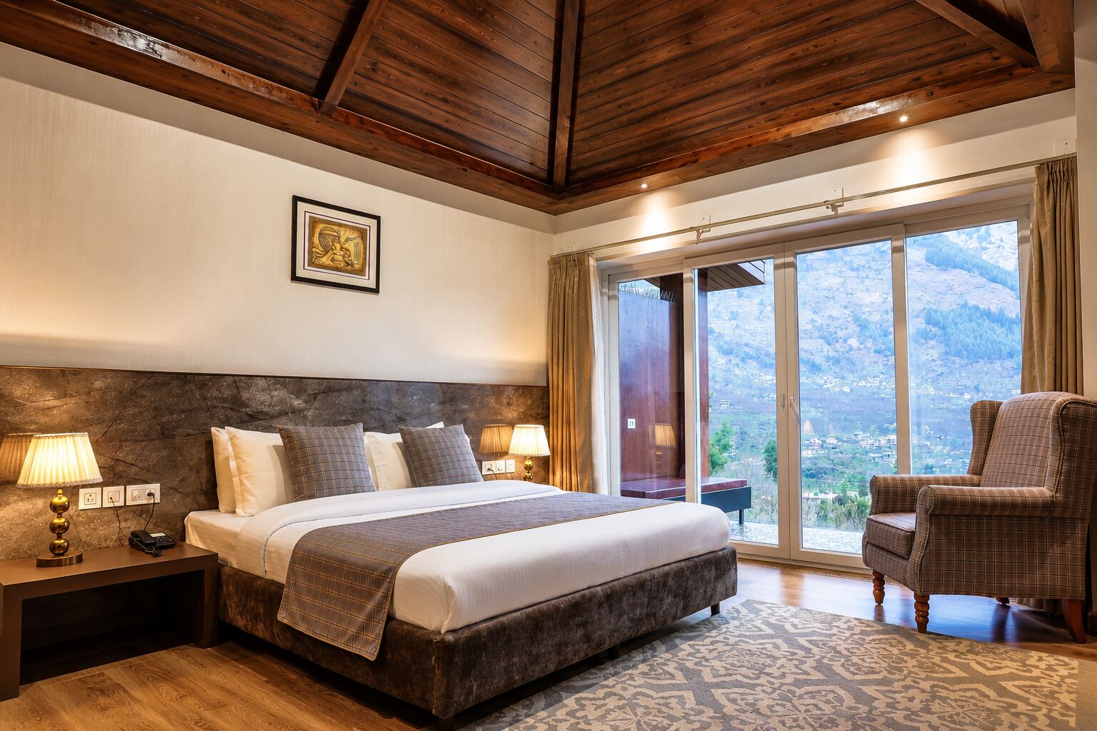 Ein Hotelzimmer mit einem großen Bett, zwei Nachttischlampen, einem Nachttisch, einem Sessel und Blick auf eine gebirgige Landschaft durch eine Glasschiebetür.