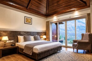 A hotel room with a large bed, two bedside lamps, a nightstand, a chair, and a view of a mountainous landscape through a sliding glass door. - The Voyage Resort, Manali (Manali)