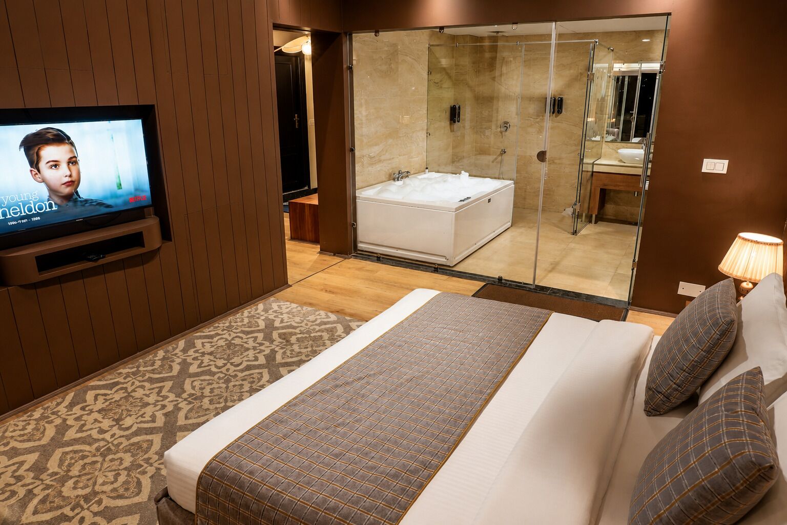 Habitación de hotel con televisor, una cama grande con colcha estampada, baño con cabina de ducha de vidrio y alfombra con diseño.