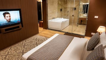 Habitación de hotel con televisor, una cama grande con colcha estampada, baño con cabina de ducha de vidrio y alfombra con diseño.